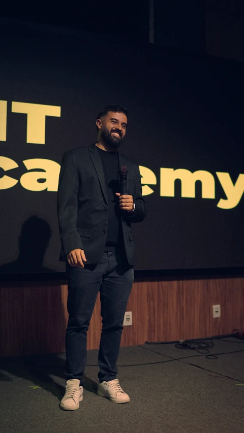 Israel Meira sorri no palco com o microfone na mão, diante do telão da IMT Academy.