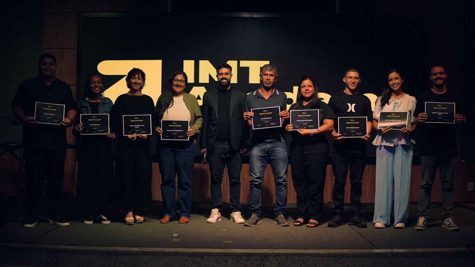 Turma de formandos do curso Cinegrafista da IMT Academy no palco, cada aluno segurando o seu certificado, com Israel Meira ao centro e o logo da IMT Academy no telão ao fundo.