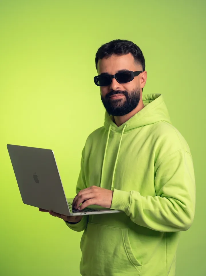Israel Meira de moletom lime e óculos escuros, digitando num MacBook aberto, em estúdio com fundo lime