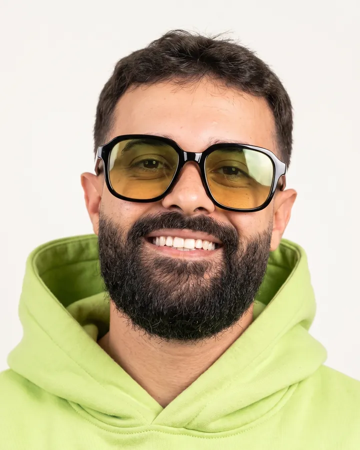 Retrato de Israel Meira sorrindo, de óculos com lente âmbar e moletom verde-limão, em fundo claro