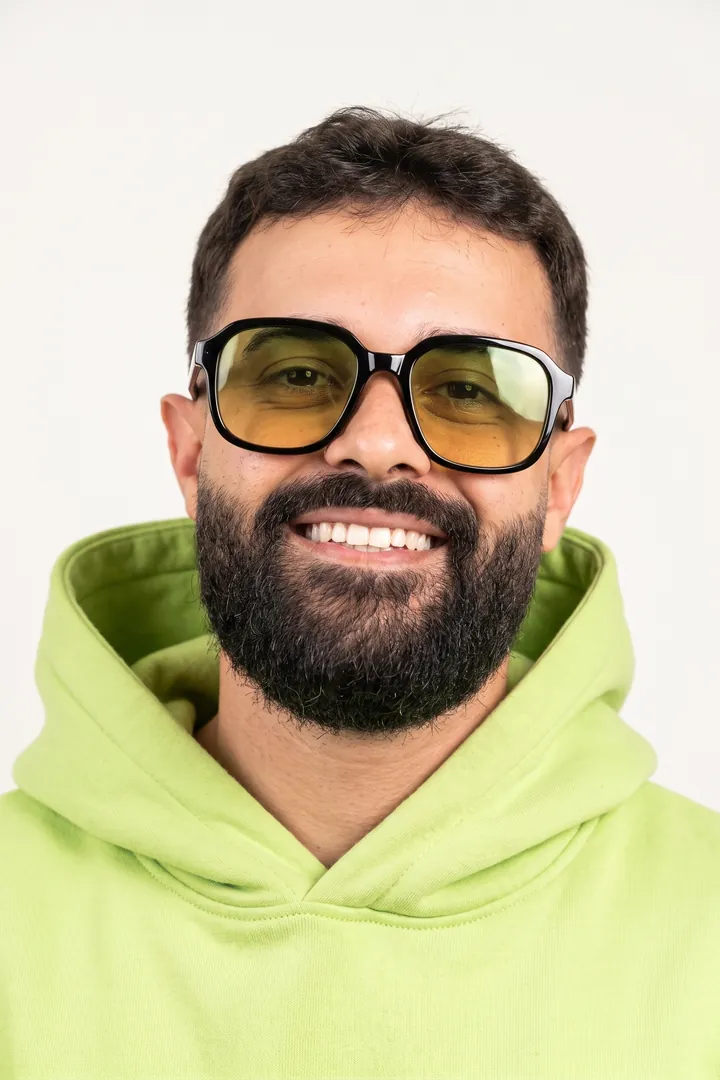 Israel Meira sorrindo, de óculos de lente amarela e moletom lime, sobre fundo claro
